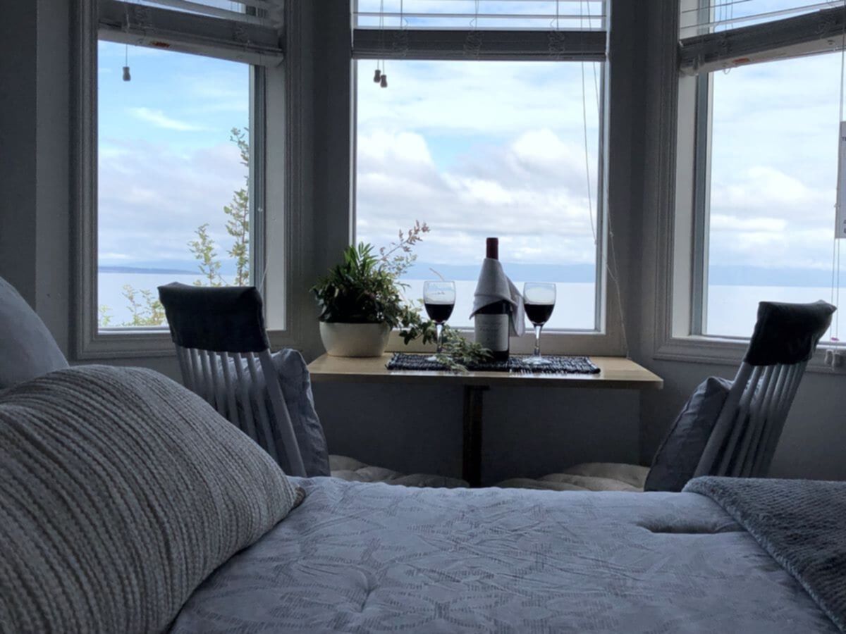 Cozy beachfront cottage interior with a queen bed, dining table set for two with wine glasses and a bottle, large windows showcasing ocean views and a relaxing atmosphere.